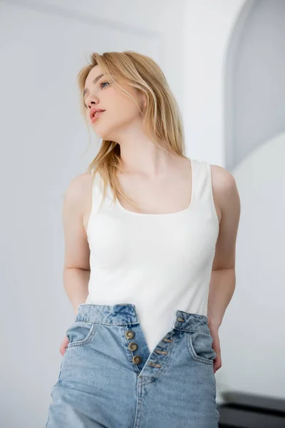 Retrato de una joven rubia en body blanco y jeans mirando a casa - foto de stock