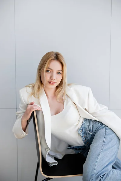 Hübsche junge Frau in Jeans und Jacke lächelt auf Stuhl — Stockfoto