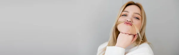 Jolie femme blonde avec des cheveux près des lèvres regardant la caméra isolée sur gris, bannière — Photo de stock