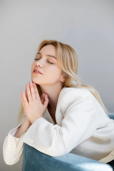 Young woman in white jacket closing eyes while sitting on chair isolated on grey — Stock Photo
