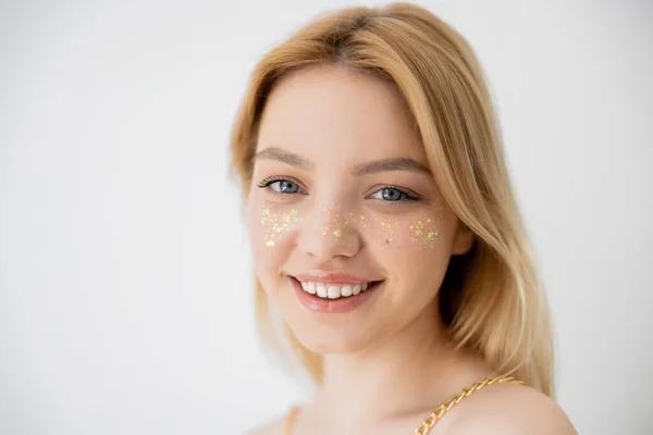 Mujer rubia alegre con maquillaje brillante mirando a la cámara aislada en gris - foto de stock