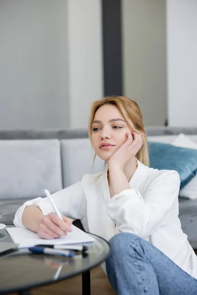 Pensive woman in blouse writing on notebook on coffee table — Stock Photo