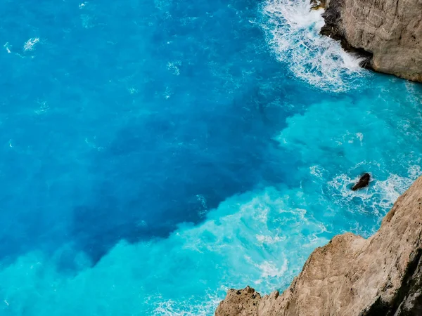 Вид сверху ярко-голубой бирюзовой морской воды с серыми скалами. Бушующие волны обрушиваются на скалы возле пляжа Закинф Греция. — стоковое фото