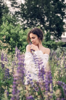 Beyaz keten gömlek ve şapka giymiş güzel bir kadın mavi ve beyaz çiçeklerle dolu bir tarlada yürüyor. Bahar. Yaz. Doğada 