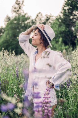 Beyaz keten gömlek ve şapka giymiş güzel bir kadın mavi ve beyaz çiçeklerle dolu bir tarlada yürüyor. Bahar. Yaz. Doğada 