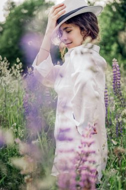 Beyaz keten gömlek ve şapka giymiş güzel bir kadın mavi ve beyaz çiçeklerle dolu bir tarlada yürüyor. Bahar. Yaz. Doğada 