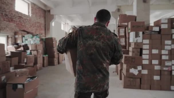 Military man carries a heavy cardboard package and puts it on the pile of boxes in the humanitarian war centre. — Vídeos de Stock
