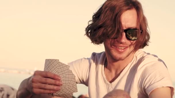 Giovane maschio con i capelli lunghi gioca a carte su una spiaggia e sorride — Video Stock