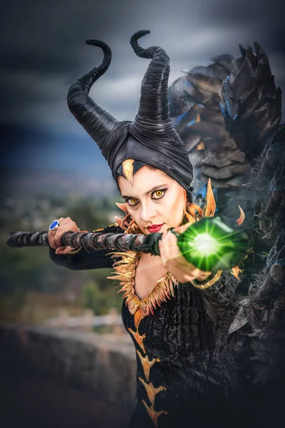MALEFICENT COSPLAY. PORTRAIT, DARK CHARACTER WITH SHARP HORNS AND STRONG WINGS. EVIL FAIRY IN BLACK DRESS. YELLOW EYES. VERTICAL.