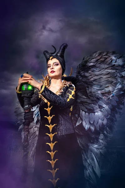 MALEFICENT COSPLAY. PORTRAIT, DARK CHARACTER WITH SHARP HORNS AND STRONG WINGS. EVIL FAIRY IN BLACK DRESS. YELLOW EYES. VERTICAL. COLD COLORS.