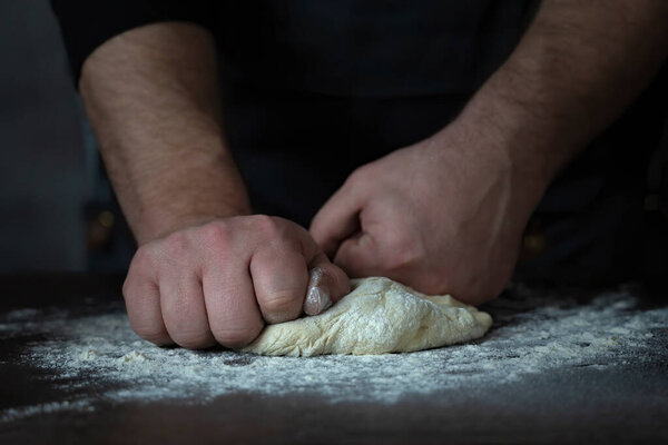 Baking fresh bread in the bakery, hands of male knead dough on kitchen table with flour
