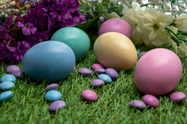 Pastel colored Easter eggs and pastel colored chocolate candy on green grass , against colored flowers background.