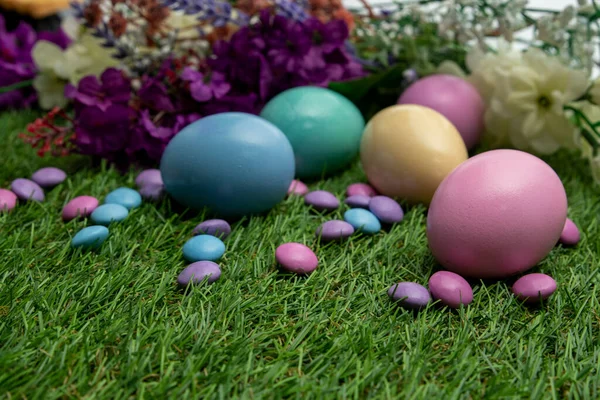 Pastel colored Easter eggs and pastel colored chocolate candy on green grass , against colored flowers background.