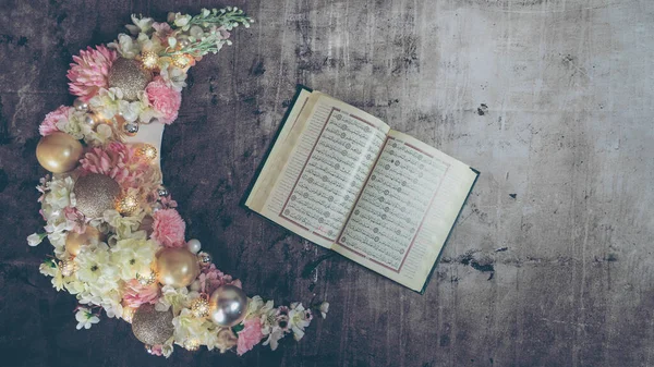 Crescent of Ramadan covered with flowers and lit up and the Holy Quran book.