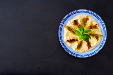 Mavi kenarlı humus kasesi, siyah ahşap arka planda bitkilerle ve zeytinyağıyla süslenmiş.. 