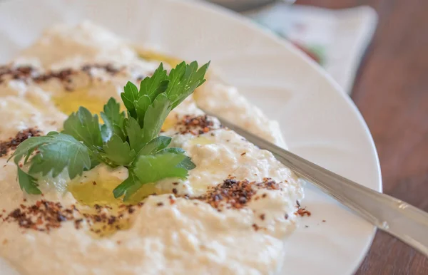 Mavi kenarlı humus kasesi, siyah ahşap arka planda bitkilerle ve zeytinyağıyla süslenmiş.. 