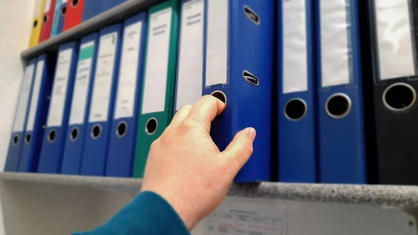 Man hand reaching for a blue office Paper folder in a row with different colors on a office shelf.