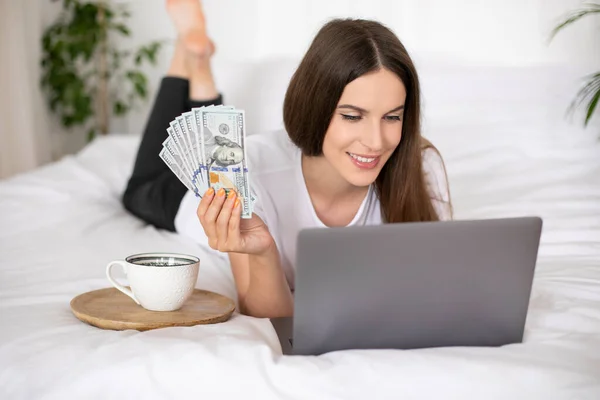 Yatakta yatan ve elinde dolar olan genç bir kız dizüstü bilgisayar kullanıyor. Evden çalışıyorum. Yüksek kalite fotoğraf