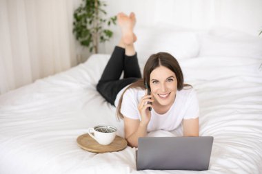 Gülümseyen esmer kız evde yatıp dizüstü bilgisayarda çalışıyor ve cep telefonuyla konuşuyor. Yüksek kalite fotoğraf