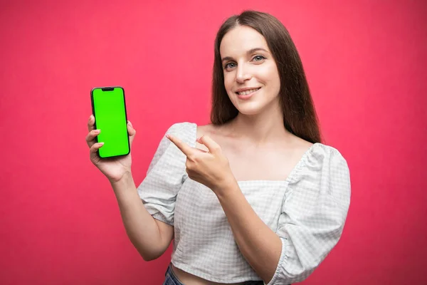 Mutlu kadın ayakta duruyor, akıllı telefonu tutuyor ve pembe arka planda telefonu işaret ediyor. Yeşil ekran telefon ekranı. Yüksek kalite fotoğraf