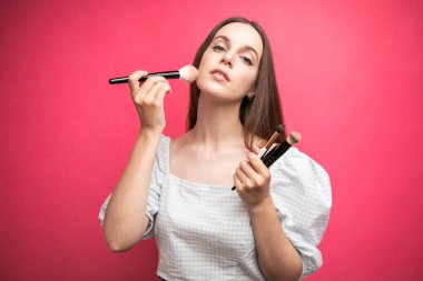 Çekici bir genç kadının portresi pembe arka planda izole bir şekilde duruyor, elinde makyaj fırçası var. Yüksek kalite fotoğraf