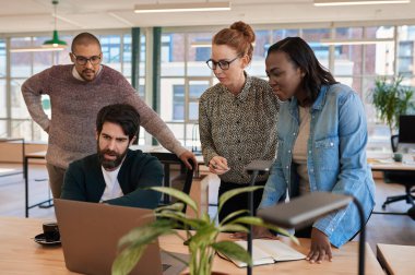 Modern bir ofisteki masanın etrafında dizüstü bilgisayarla çalışan çeşitli odaklanmış genç iş adamları.