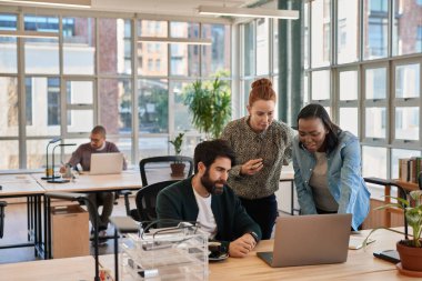 Modern bir ofiste masa başında çalışırken dizüstü bilgisayarla konuşan bir grup genç iş adamı.
