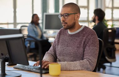 Geniş bir açık ofis alanında bilgisayar üzerinde çalışan odaklanmış genç işadamı, arka planda iş arkadaşları konuşuyor.