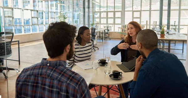 Young businesswoman explaining something to a diverse group of colleagues during a meeting over coffee in a modern office space