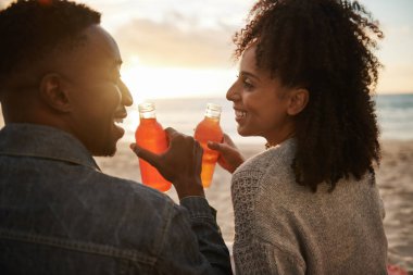 Rear view of a smiling young multiethnic couple drinking juice while sitting on a sandy beach at sunset