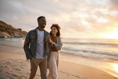 Loving young multiethnic couple smiling and talking together while walking arm in arm along a sandy beach at sunset