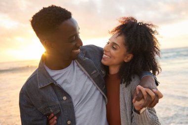 Loving young multiethnic couple smiling at each while standing arm in arm together on a beach at sunset