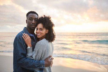 Portrait of a loving young multiethnic couple standing together in each other's arms on a sandy beach at sunset