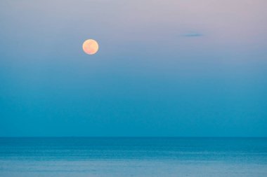 Dolunay rengarenk bir günbatımında denizin üzerinde yükseliyor.