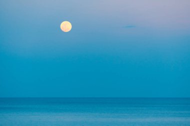 Dolunay rengarenk bir günbatımında denizin üzerinde yükseliyor.