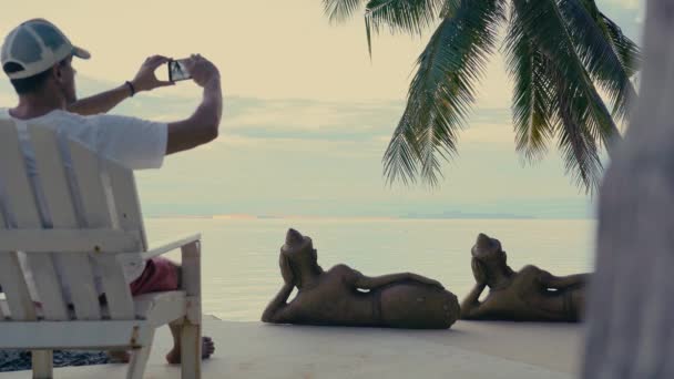 日落时 一个男人坐在海边棕榈树下的木椅上 用智能手机拍照 — 图库视频影像