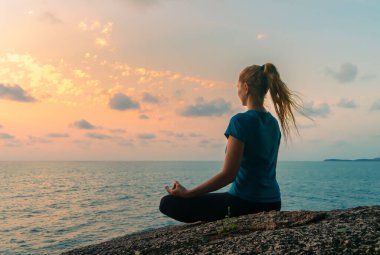 Lotus pozisyonundaki genç mutlu kadın gün doğumunda deniz kenarındaki kayalarda meditasyon yapıyor. Arkaplanda renkli bir gökyüzü.