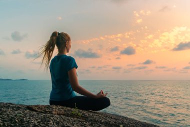 Lotus pozisyonundaki genç mutlu kadın gün doğumunda deniz kenarındaki kayalarda meditasyon yapıyor. Arkaplanda renkli bir gökyüzü.