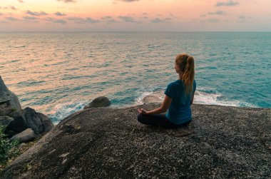 Lotus pozisyonundaki genç bir kadın gün doğumunda deniz kenarındaki kayalar üzerinde meditasyon yapar. Arkaplanda renkli bir gökyüzü.