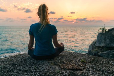 Lotus pozisyonundaki genç bir kadın gün doğumunda deniz kenarındaki kayalar üzerinde meditasyon yapar. Arkaplanda renkli bir gökyüzü.