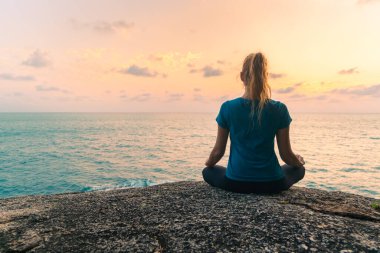 Lotus pozisyonundaki genç bir kadın gün doğumunda deniz kenarındaki kayalar üzerinde meditasyon yapar. Arkaplanda renkli bir gökyüzü.