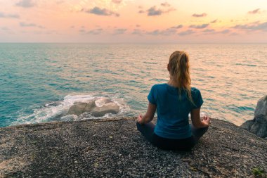 Lotus pozisyonundaki genç bir kadın gün doğumunda deniz kenarındaki kayalar üzerinde meditasyon yapar. Arkaplanda renkli bir gökyüzü.