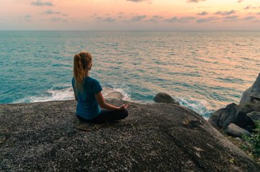 Lotus pozisyonundaki genç bir kadın gün doğumunda deniz kenarındaki kayalar üzerinde meditasyon yapar. Arkaplanda renkli bir gökyüzü.