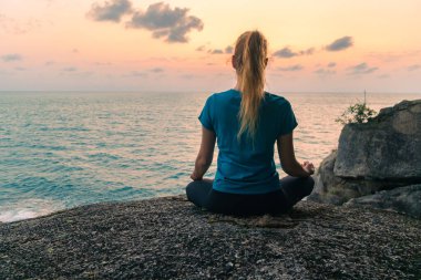 Lotus pozisyonundaki genç bir kadın gün doğumunda deniz kenarındaki kayalar üzerinde meditasyon yapar. Arkaplanda renkli bir gökyüzü.