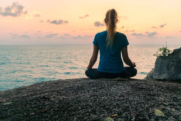 Lotus pozisyonundaki genç bir kadın gün doğumunda deniz kenarındaki kayalar üzerinde meditasyon yapar. Arkaplanda renkli bir gökyüzü.