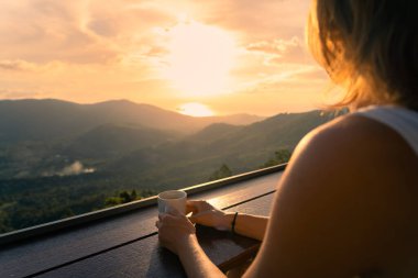Beyaz elbiseli genç, ince bir kadın bir kafede bir fincan kahveyle tek başına oturuyor ve vadide renkli günbatımının tadını çıkarıyor. Arka planda deniz ve dağlarda.