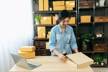 Kadın paket kutusunu masaya koydu. Müşteriye mal teslimatını kontrol ediyor. Asyalı bir kadın ofis masasında küçük bir iş kuruyor. Girişimci Asyalı kadın teslimat için ürün topluyor
