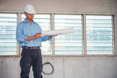 İnşaat mühendisi planı tutuyor. İnşaat alanında çalışan işçilerden mavi gömlekli, güvenlik önleyici beyaz şapka giyiyor. Mimarlık mühendisi, inşaat mühendisi, emlak konseptine bakıyor.