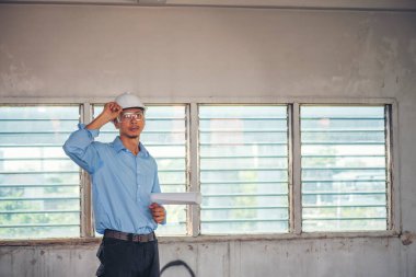 İnşaat mühendisi planı tutuyor. İnşaat alanında çalışan işçilerden mavi gömlekli, güvenlik önleyici beyaz şapka giyiyor. Mimarlık mühendisi, inşaat mühendisi, emlak konseptine bakıyor.