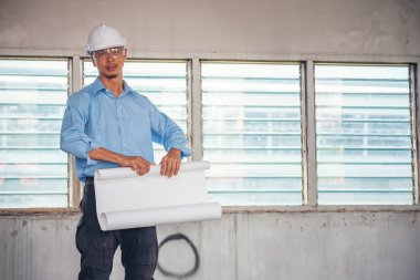 İnşaat mühendisi planı tutuyor. İnşaat alanında çalışan işçilerden mavi gömlekli, güvenlik önleyici beyaz şapka giyiyor. Mimarlık mühendisi, inşaat mühendisi, emlak konseptine bakıyor.